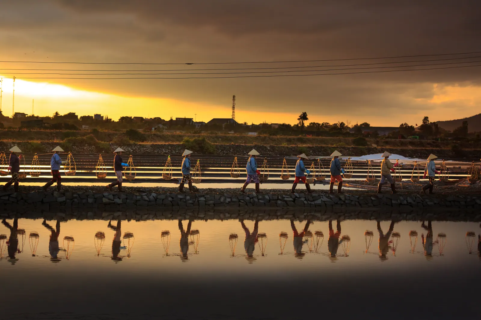 Salt workers carrying baskets along a dyke through the Hon Khoi salt fields at sunset, reflected in the flooded pans.