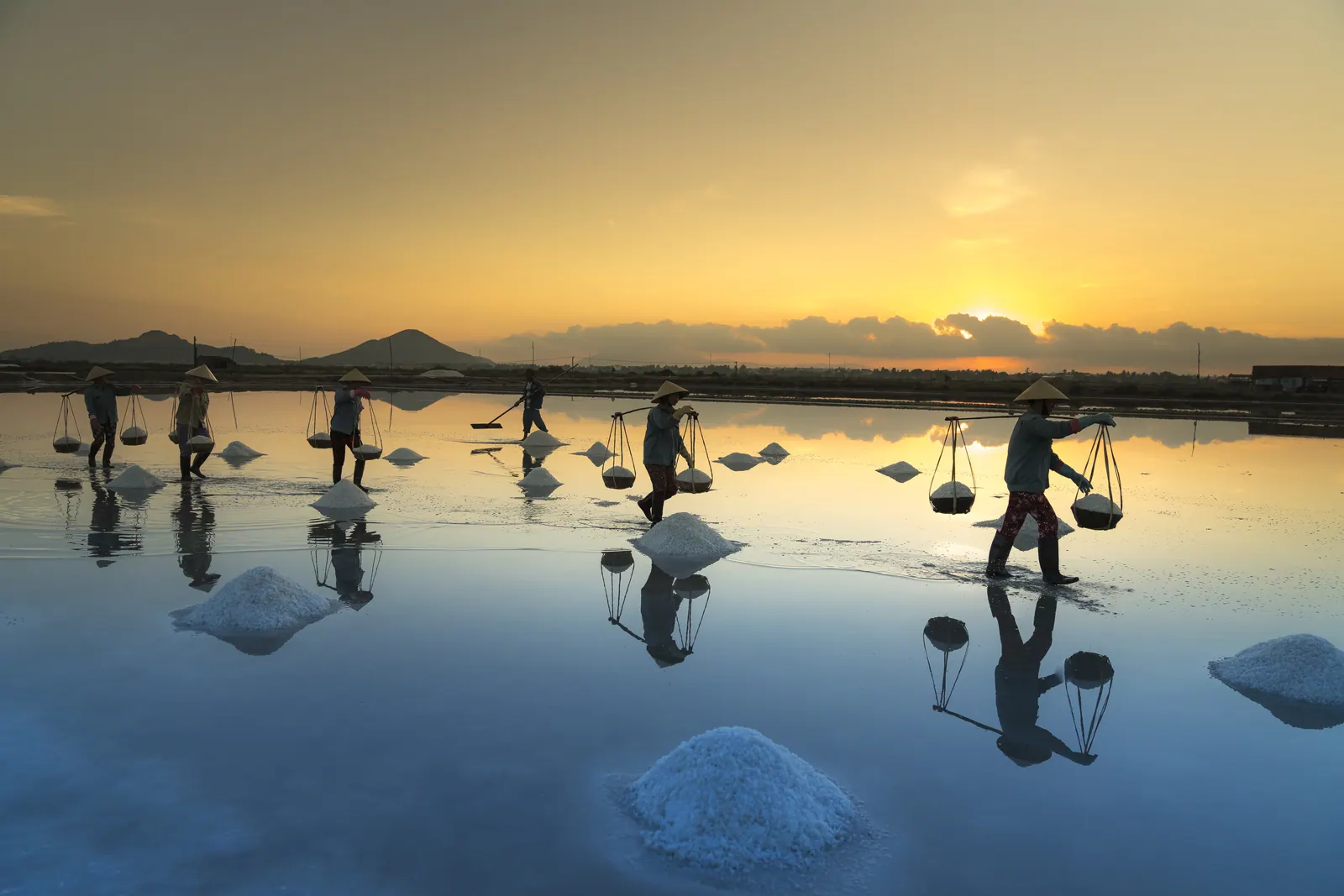 A line of salt workers with shoulder poles crossing the flooded Hon Khoi salt pans in the blue light of early morning