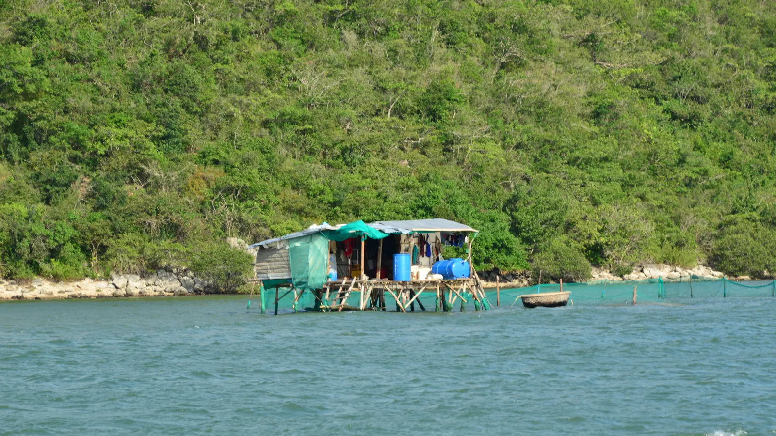 A floating fisherman's hut on the calm water of Ninh Van bay below a green hillside